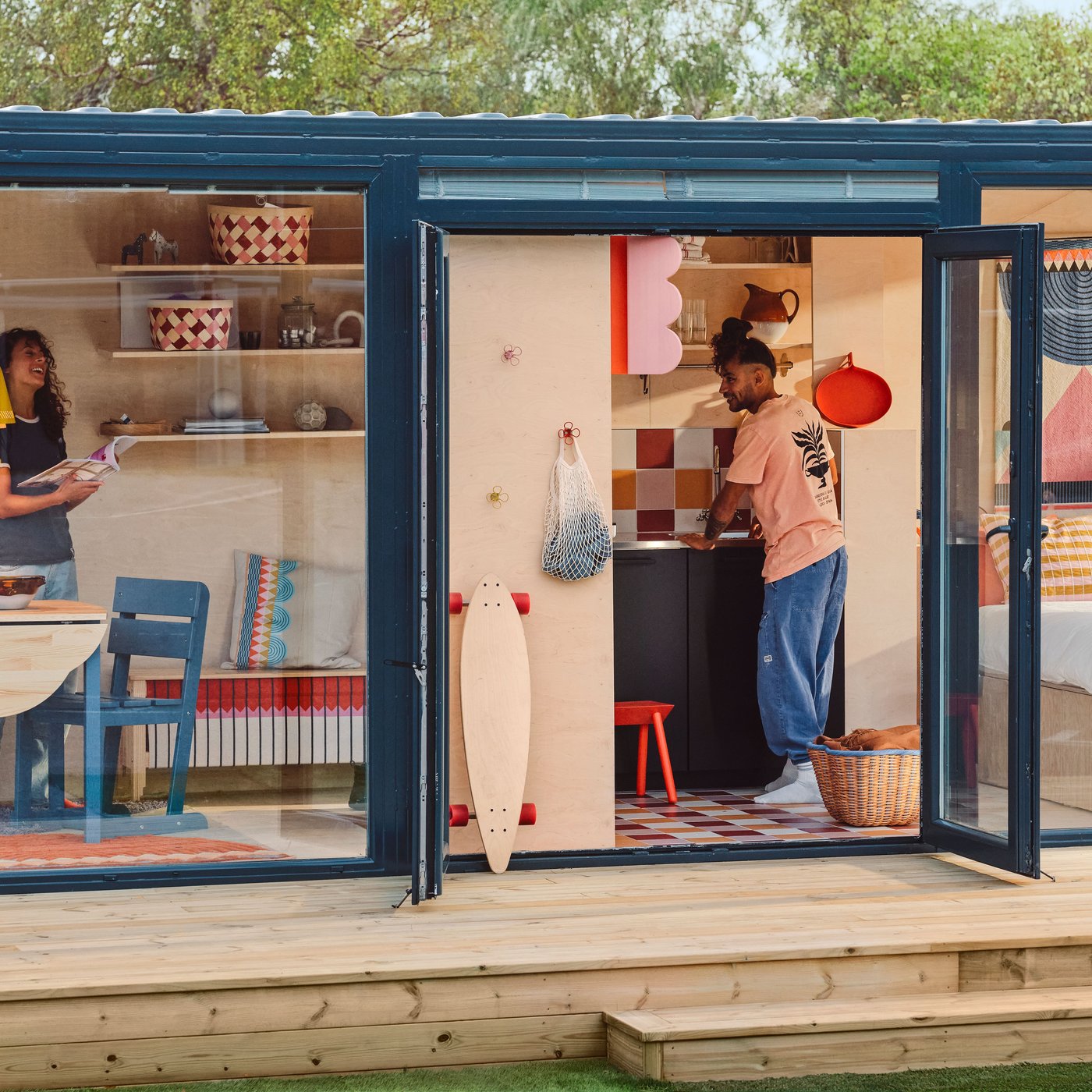 A tiny house on a deck, with glass walls and doors, seen from outside. There are OMMJÄNGE furnishings and decorations inside.