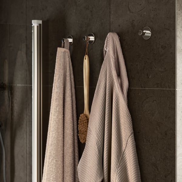 A beige, waffle-textured BJÄLVEN bathrobe hangs on a hook on the wall beside a beige VALLASÅN towel and a wooden body brush.