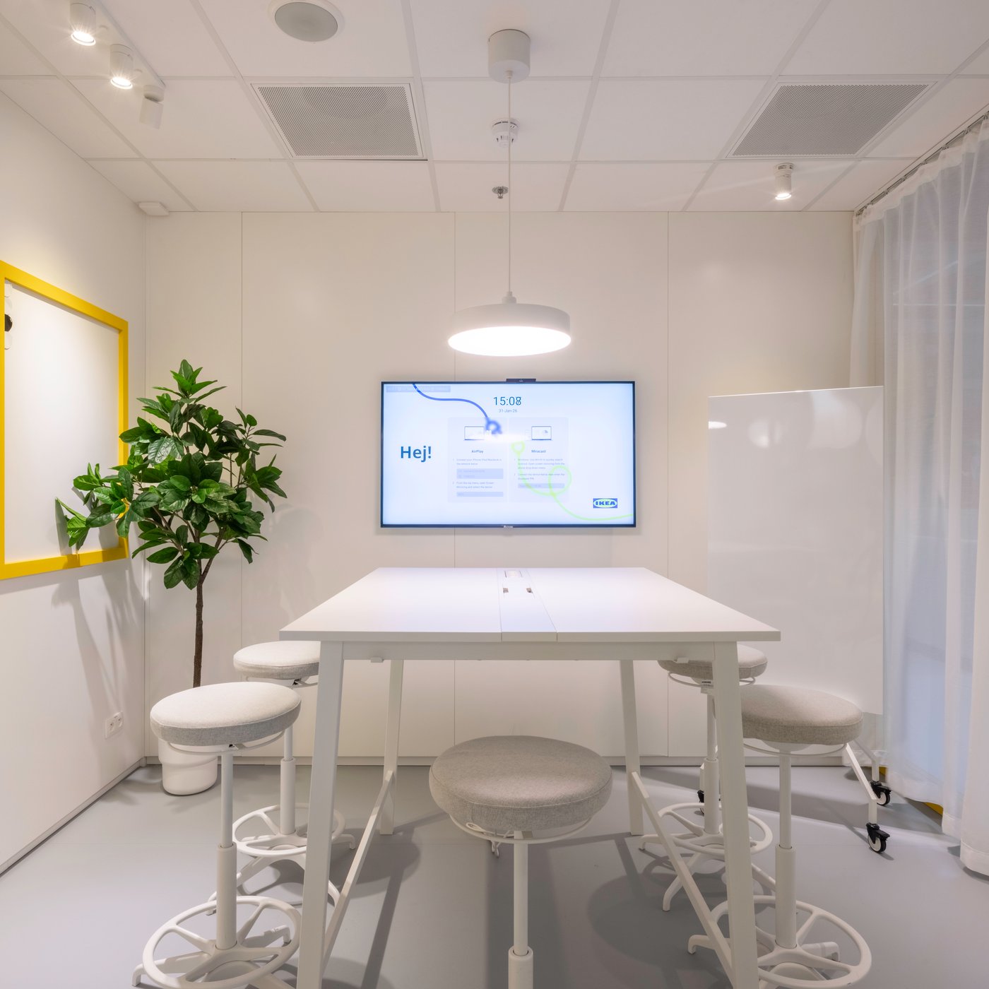 Salle de réunion blanche épurée avec des tabourets ronds, un écran de présentation affichant « Hej ! », un tableau blanc encadré de jaune et une plante verte d’intérieur.