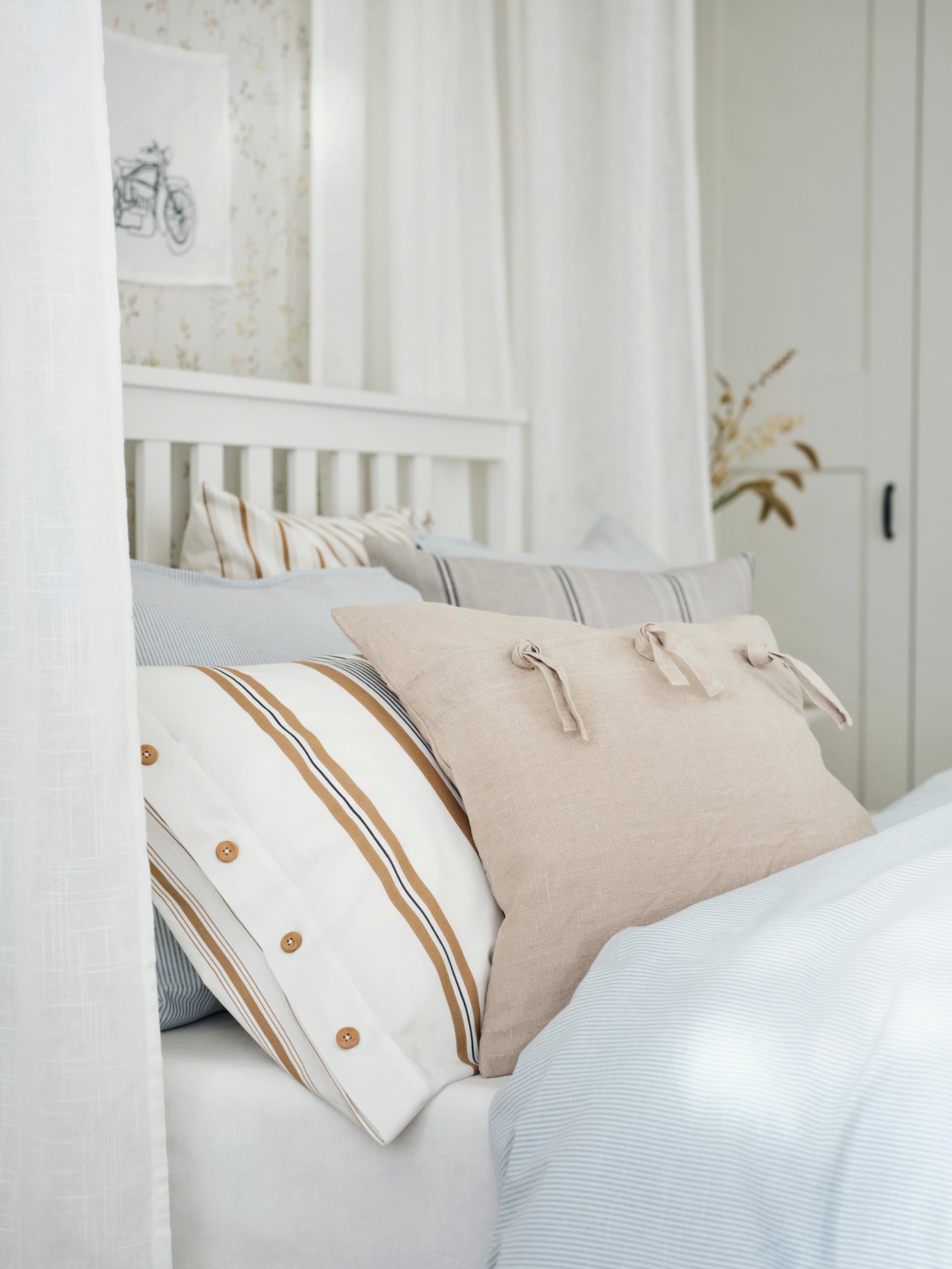 A HEMNES bed surrounded by sheer curtains with blue bed linen and cushions covered in STORTIMJAN and AINA covers on top.