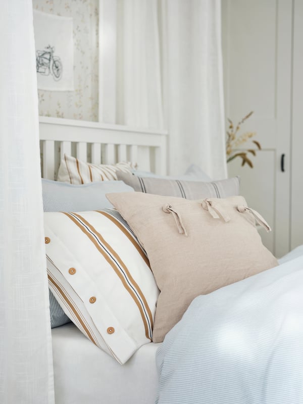 A HEMNES bed surrounded by sheer curtains with blue bed linen and cushions covered in STORTIMJAN and AINA covers on top.