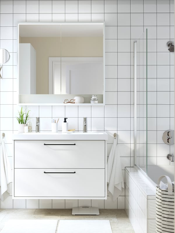 Un meuble lavabo HAVBÄCK blanc à tiroirs est fixé sur un mur de salle de bains carrelé blanc, sous une armoire à pharmacie et près d’une douche.