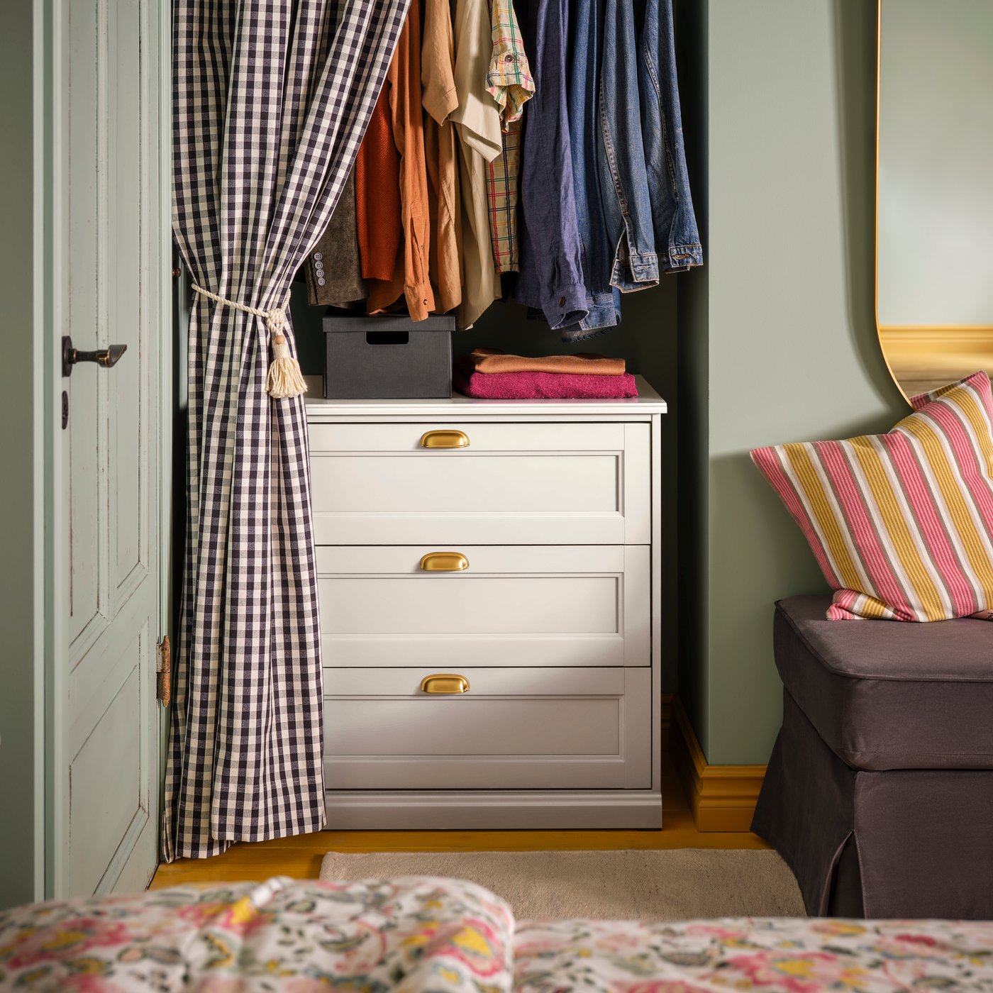 A bedroom with a FÅGELFJÄLLET chest of 3 drawers inside an alcove with clothes hanging above it, behind a checked curtain.