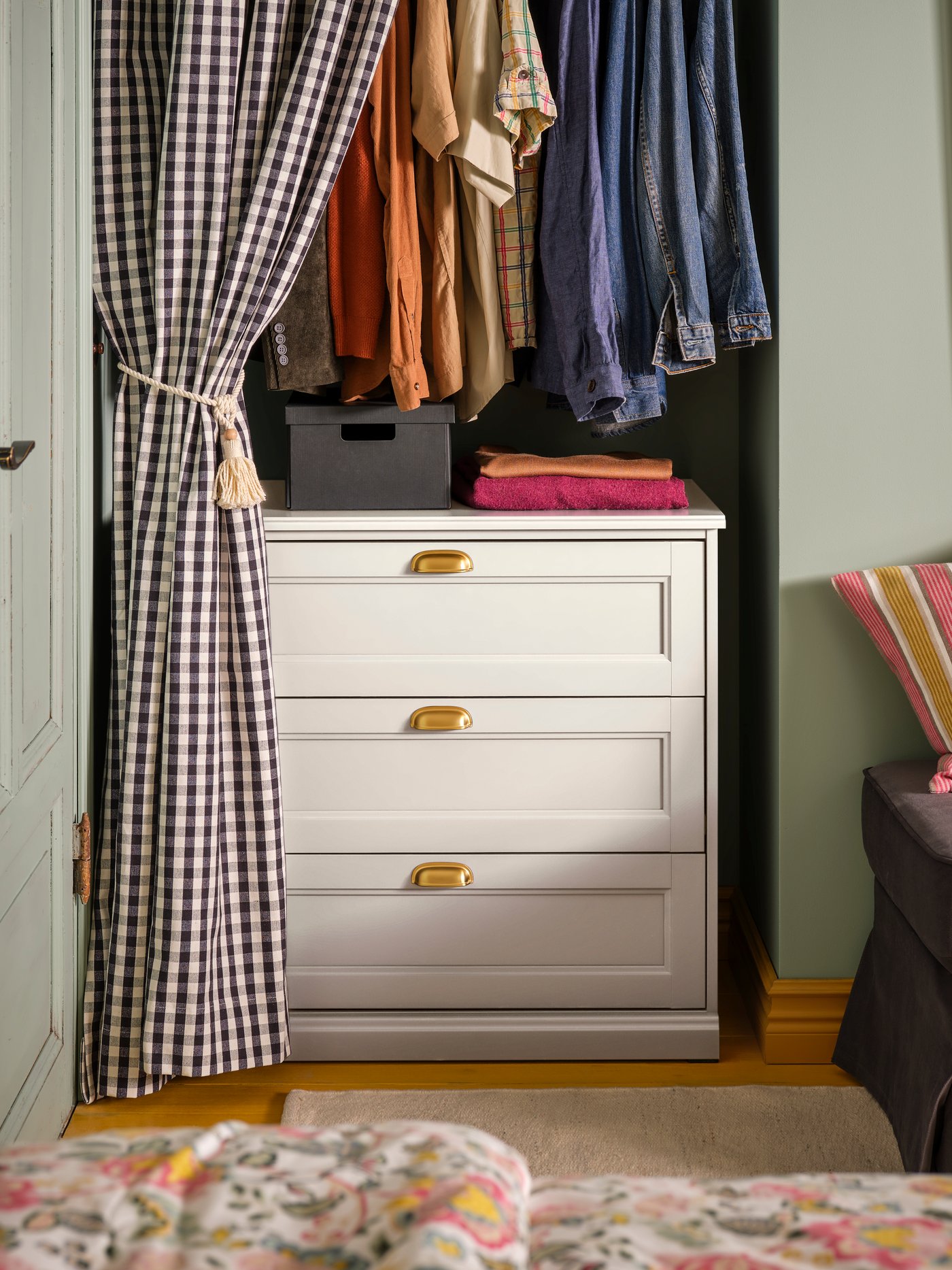 A bedroom with a FÅGELFJÄLLET chest of 3 drawers inside an alcove with clothes hanging above it, behind a checked curtain.
