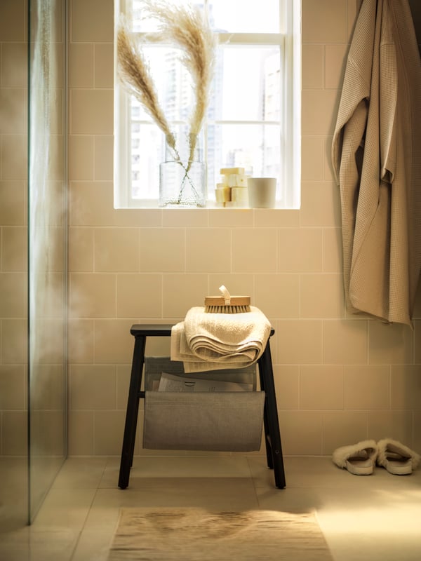 Kupatilo s krem pločicama s crnom stoličicom za odlaganje ispod prozora s travom u talasastoj staklenoj vazi.