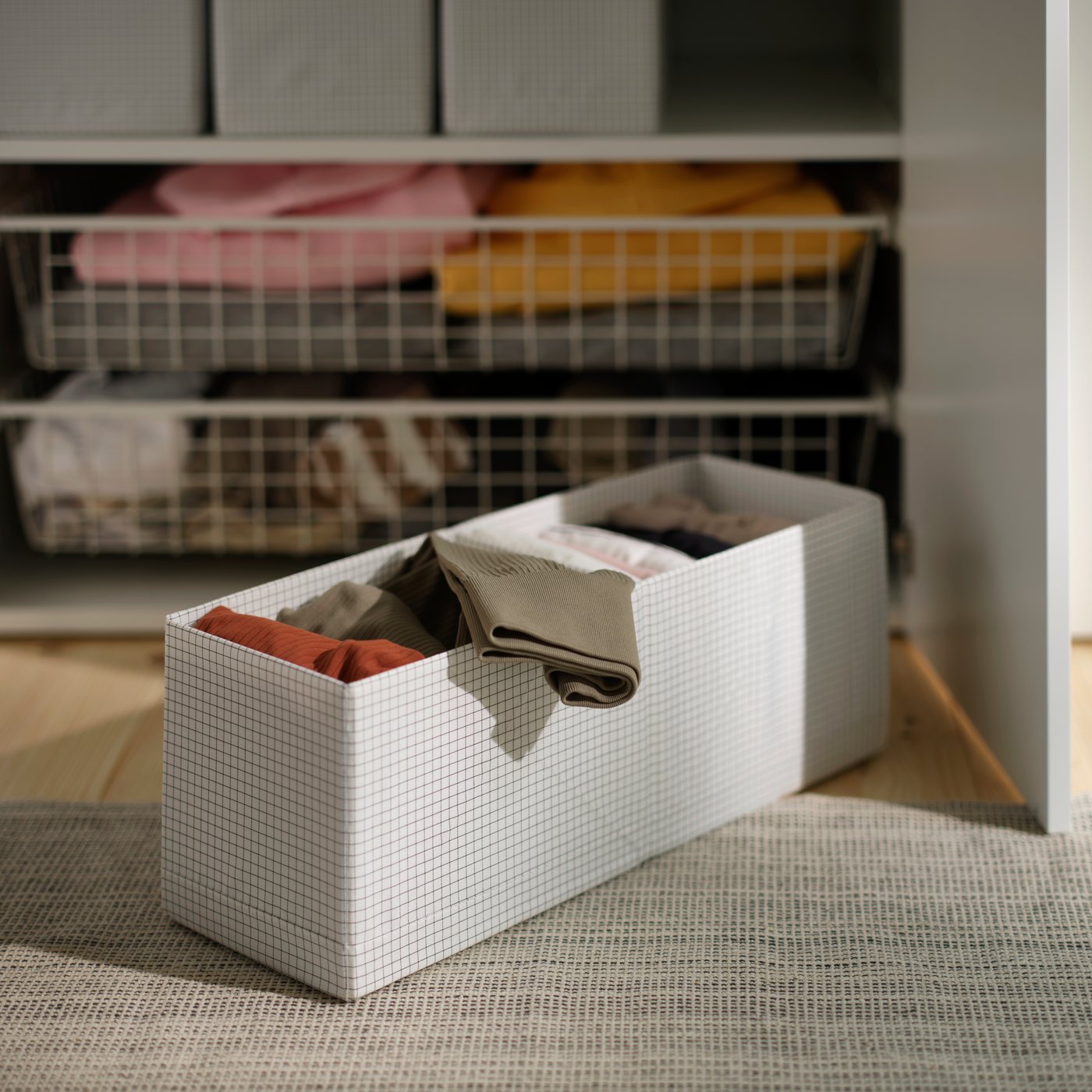 Una caja con compartimentos STUK blanca llena de ropa doblada, sobre un tapete y junto a un clóset.