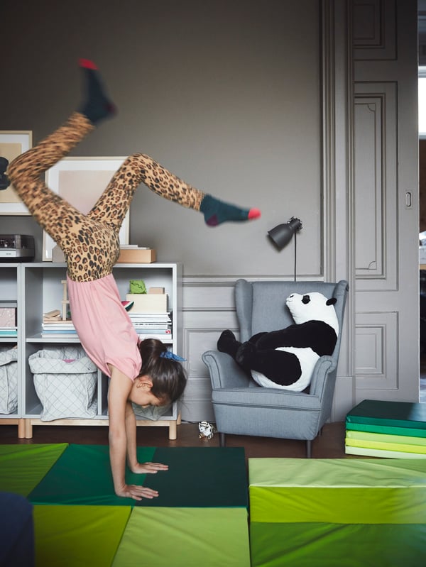 Una niña haciendo una invertida al interior de una casa, apoyada sobre colchonetas PLUFSIG verdes. Frente al muro del fondo hay un sillón STRANDMON de tamaño infantil y módulos de organización EKET. 