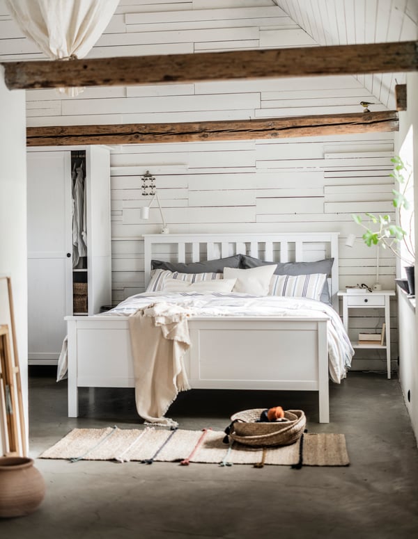 Ein Schlafzimmer im skandinavischen Stil mit Boho Akzenten wie geflochtenen Teppichen, Plaids und Pflanzen. Die weissen Schlafzimmermöbel werden ergänzt durch Textilien in natürlichen Beige und Braun-tönen.