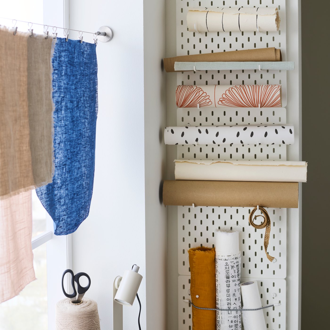 Drie witte SKÅDIS pegboards hangen verticaal aan een muur. Ze zijn uitgerust met haken en een elastisch koord waarmee rollen papier aan het ophangbord zijn vastgemaakt.