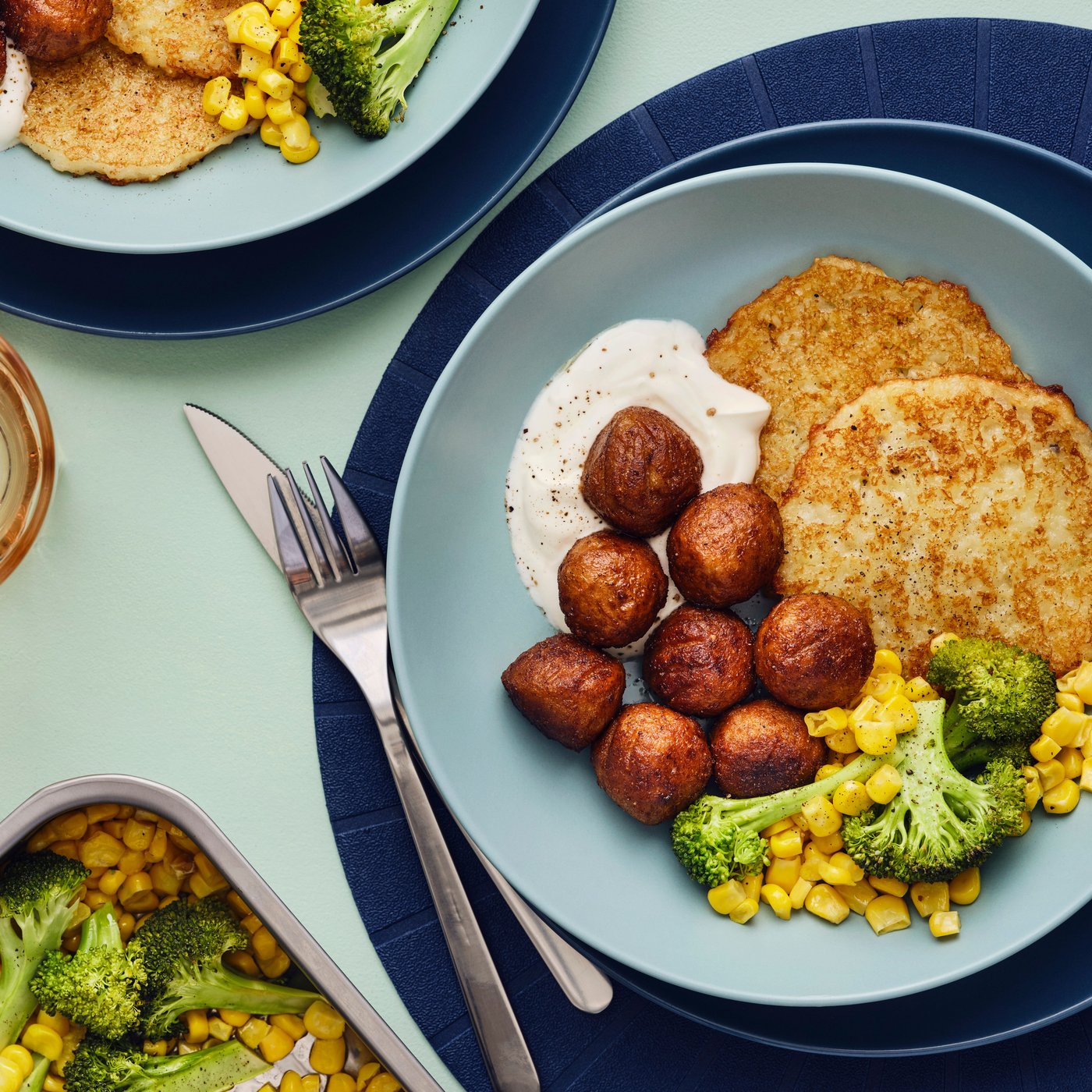 Two blue bowls filled with a meal of HUVUDROLL plant balls, potato pancakes and a mix of corn and broccoli are on a table.