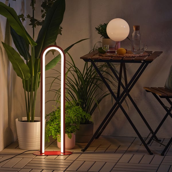 Eine dimmbare rote SOLVINDEN Stehleuchte sorgt in der Dunkelheit für eine gemütliche Atmosphäre, umgeben von grünen Pflanzen und einem Balkontisch.