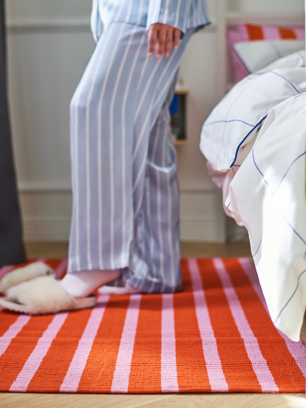 Krupni plan VÄGSKYLT tepiha prostrtog pored kreveta, na kom je osoba u pidžami koja obuva papuče.