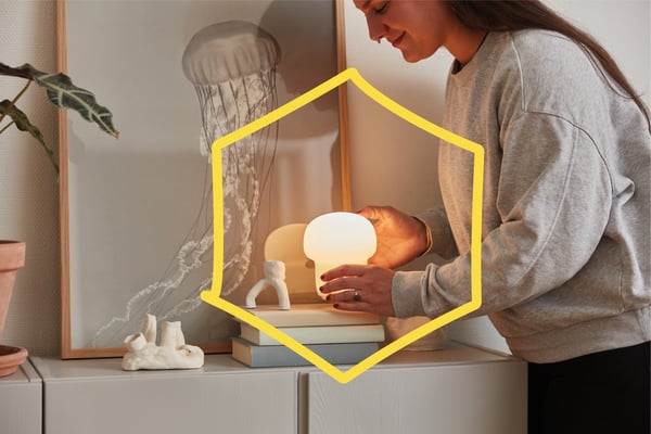 Eine Frau hält die Tischlampe TOKABO über einen Stapel Bücher auf einem weissen Schrank, hinter den Büchern ist ein gerahmtes Bild einer Qualle angebracht.