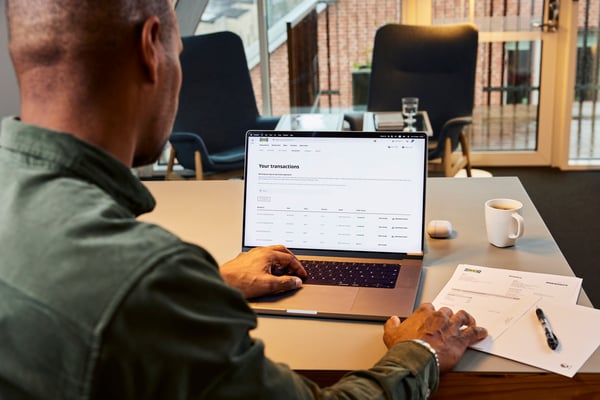 Person sitter vid ett konferensbord och använder en bärbar dator för att granska transaktioner, med dokument och kaffekopp bredvid.