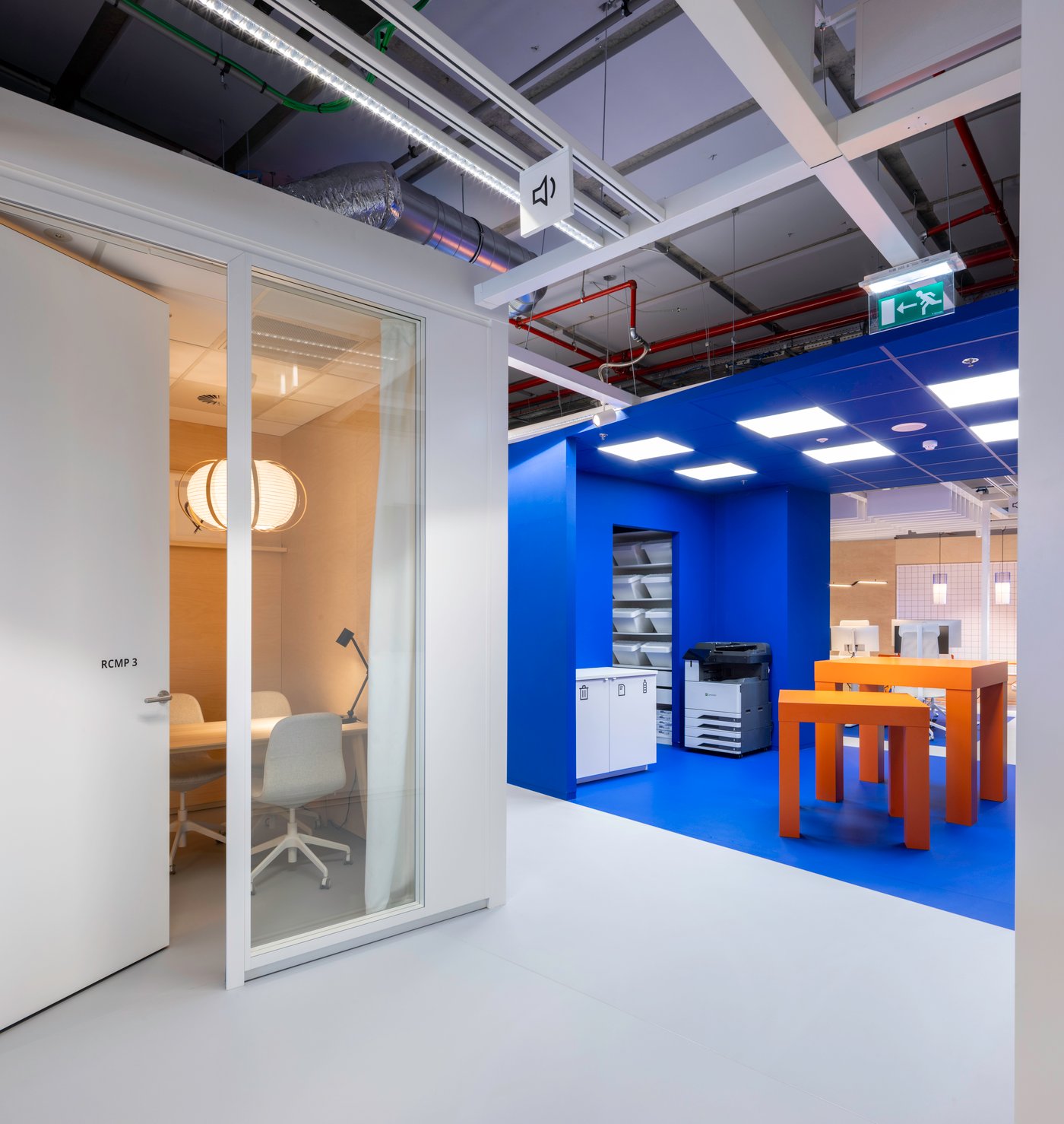 Un espace de travail bleu frais avec des tables IKEA et des touches d’orange offre une zone pratique pour l’impression et le travail de projet, juste à côté d’une salle de réunion lumineuse à l’éclairage doux et chaleureux.