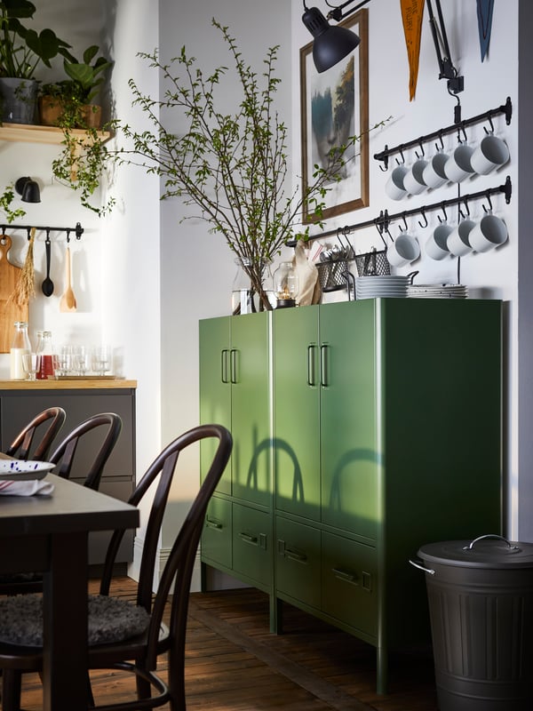 Due mobili con ante e cassetti IDÅSEN verde scuro sono appoggiati a una parete della zona pranzo. Sopra, ci sono molte tazze appese a dei binari.