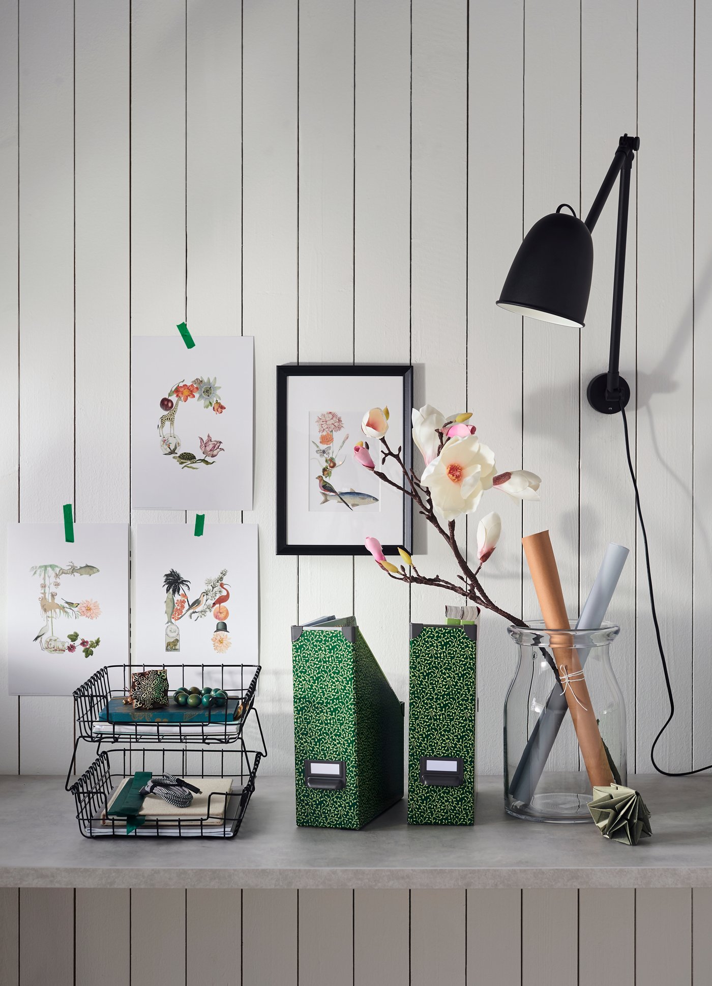 Kreativer Arbeitsplatz mit floralen Drucken an der weissen Wand, Ordnern, einer Vase mit Zweigen, gerolltem Papier und einer schwarzen Wandleuchte.