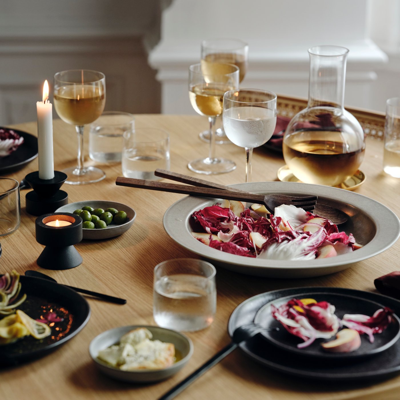 Prostřený stůl na elegantní večeři s černými svícny IKEA, zapálenou svíčkou, sklenicemi vína a vody, karafou, miskami s olivami a talíři se salátem z červené čekanky.