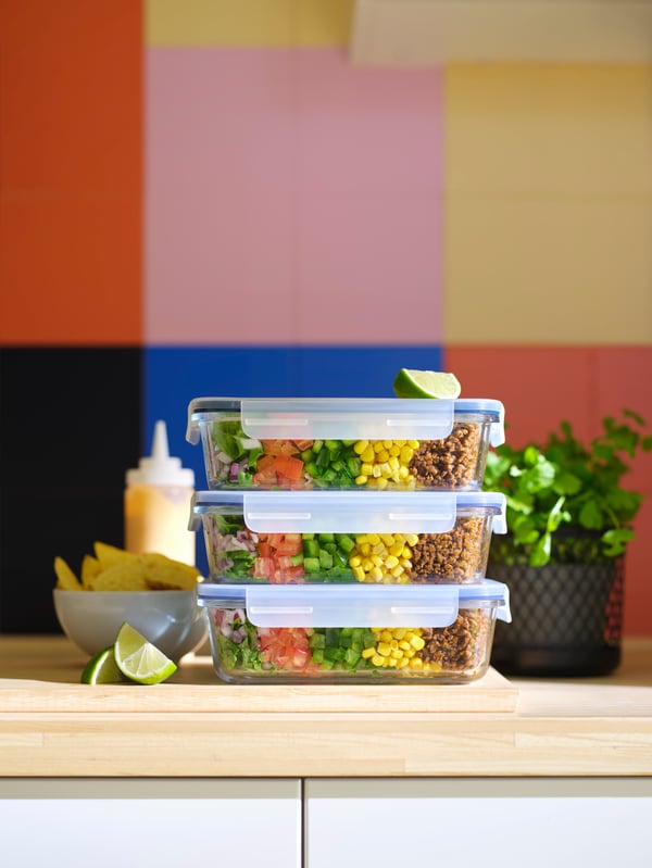 Three stacked IKEA 365+ food containers with layered salad ingredients are placed on a countertop with a colourful wall.