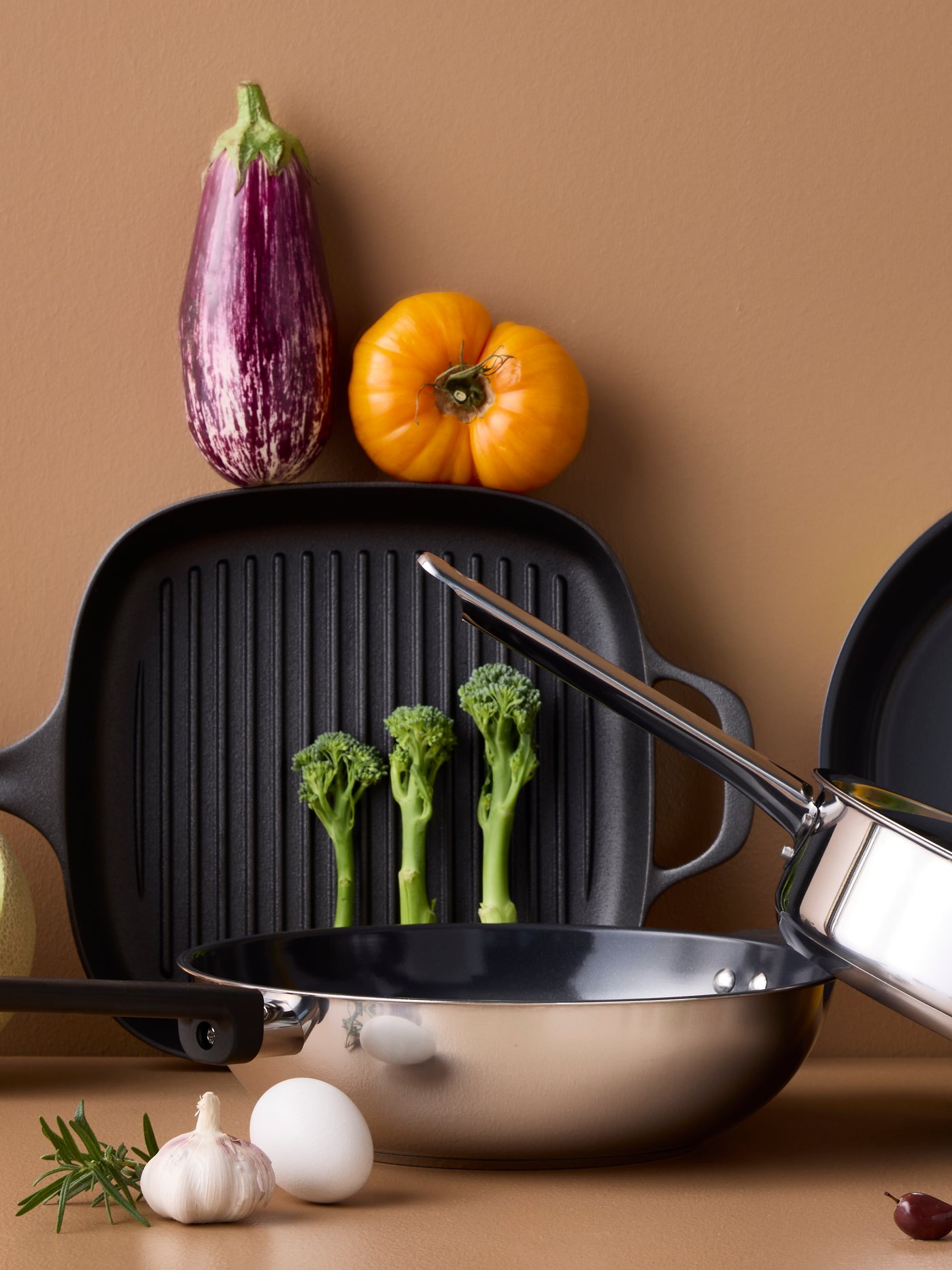 Four pans of different styles and materials are placed on a brown surface with different kinds of vegetables around them.