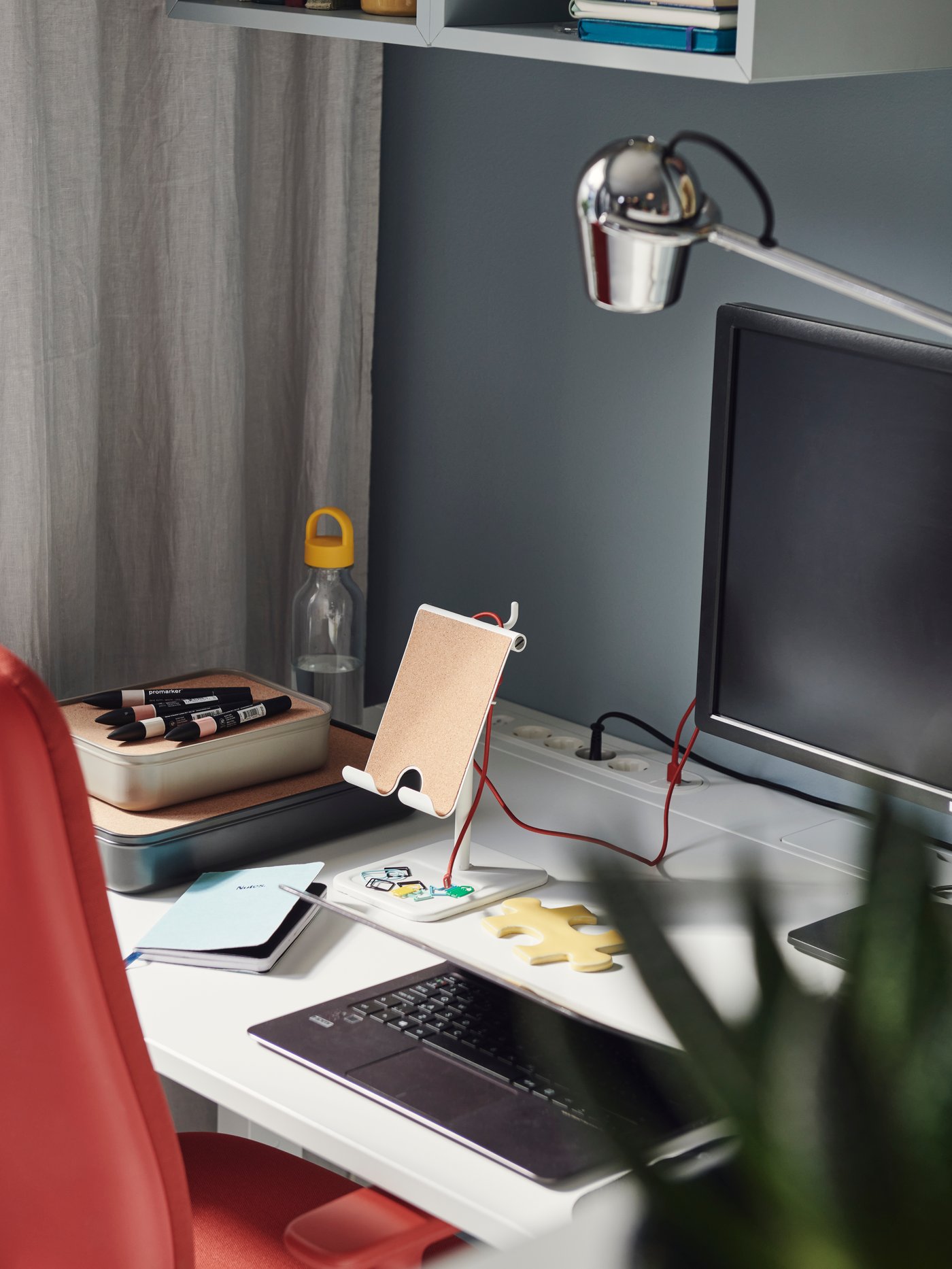 A SMÖRKULL office chair in Gräsnäs red next to a white desk, which is against a blue wall and beige curtain.
