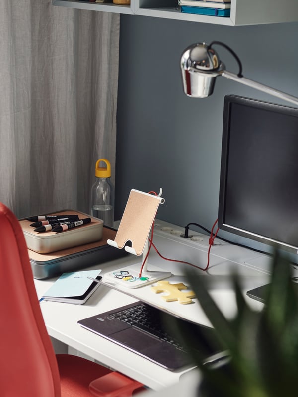A SMÖRKULL office chair in Gräsnäs red next to a white desk, which is against a blue wall and beige curtain.