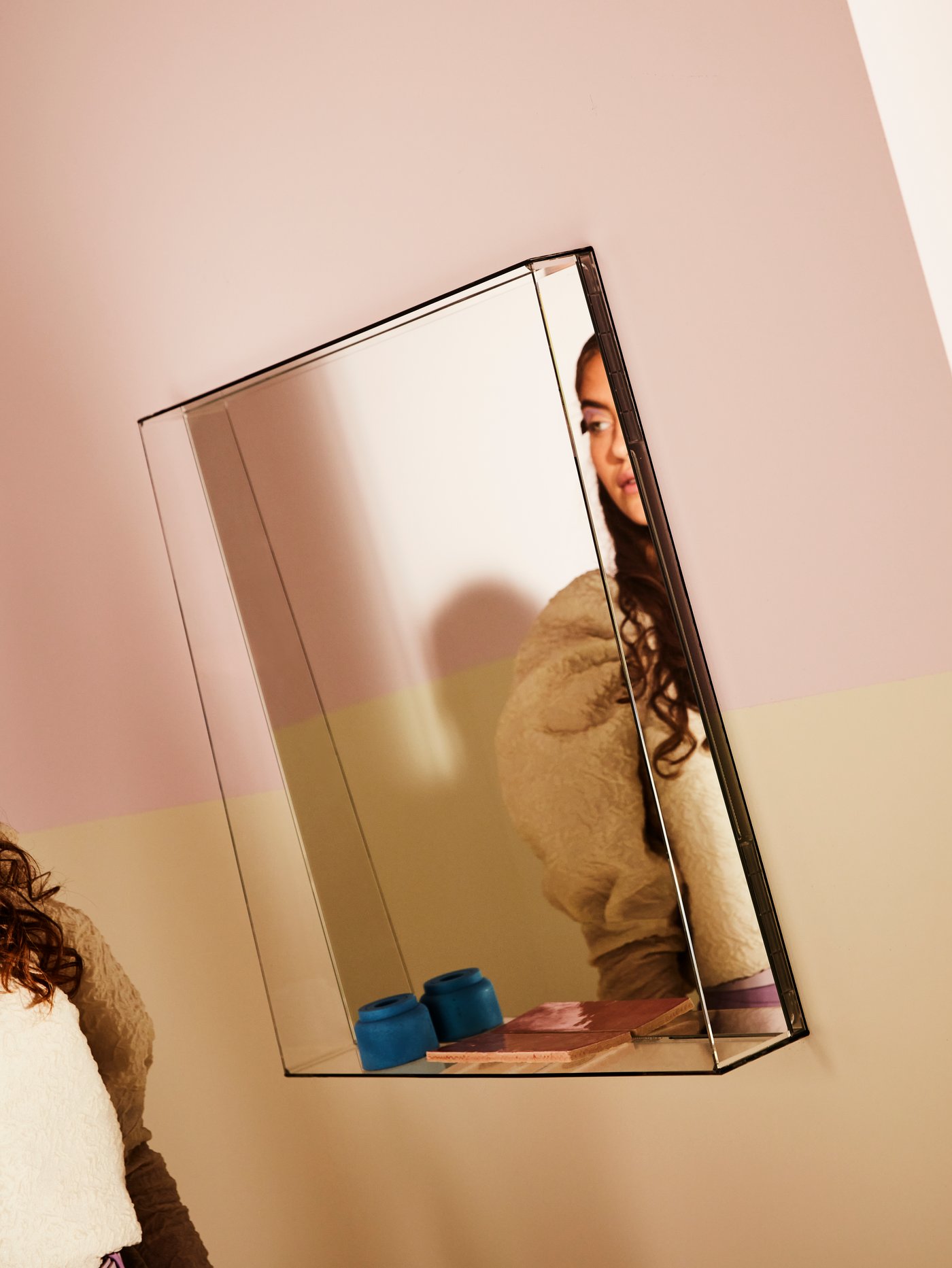 GRIMSTORP mirror with a frame acting as a shelf placed on a pastel pink and yellow wall, reflecting parts of a woman.