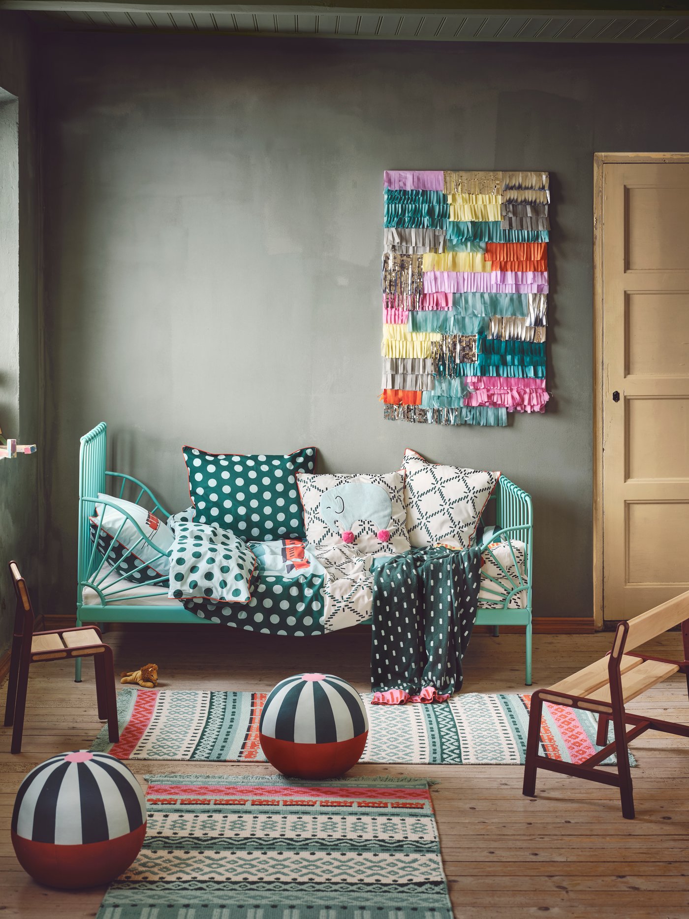Ein verspieltes Kinderzimmer im Boho-Stil mit einem grünen Bett an der Rückwand und vielen Textilien darauf. Kleine Teppiche und Kuscheltiere liegen auf dem Boden.