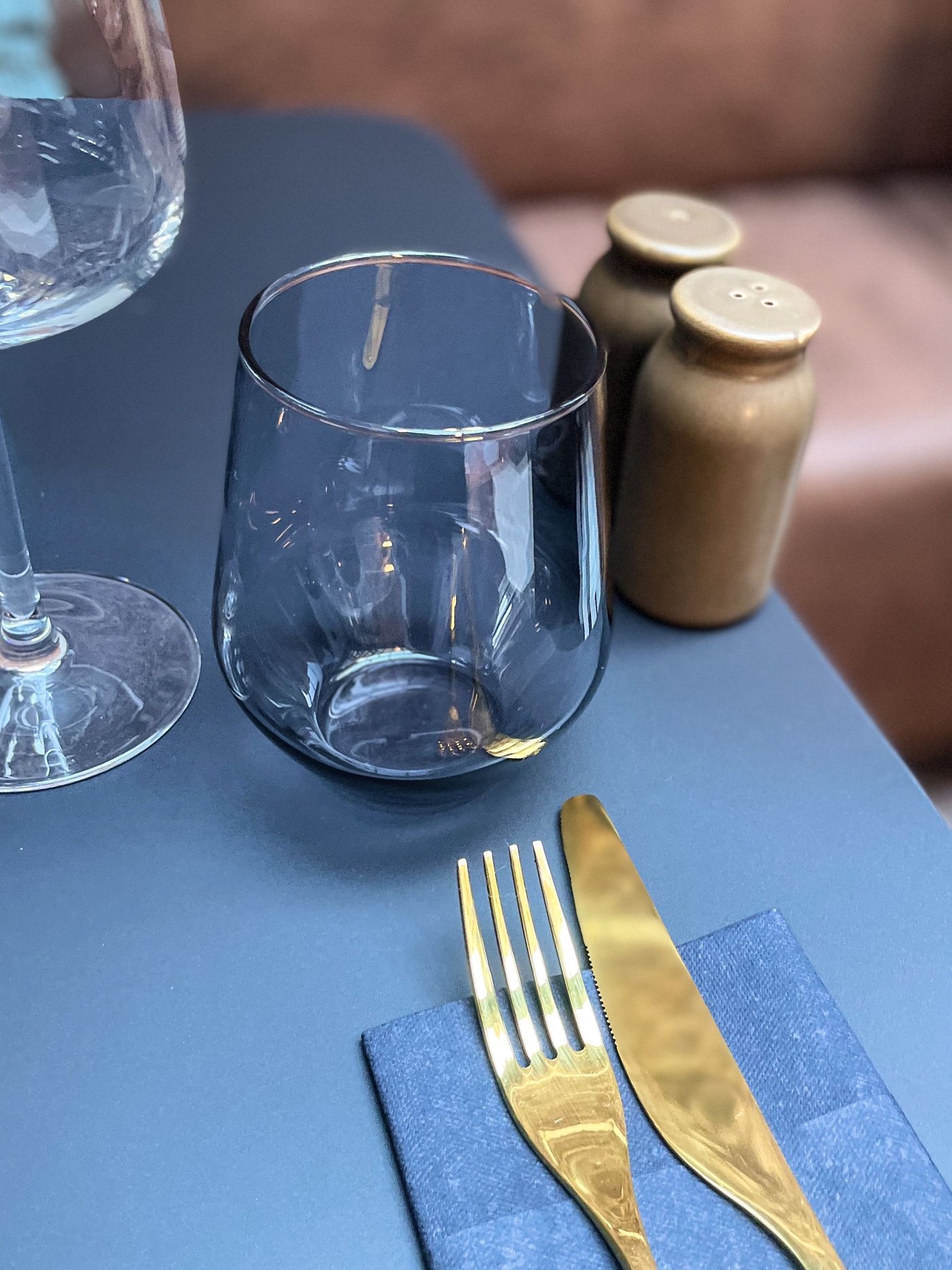 Donkerblauwe tafel met gouden bestek, glazen en bruine kruidenpotjes. Achteraan een bruine zetel, sfeervol gedekt in modern restaurantinterieur.