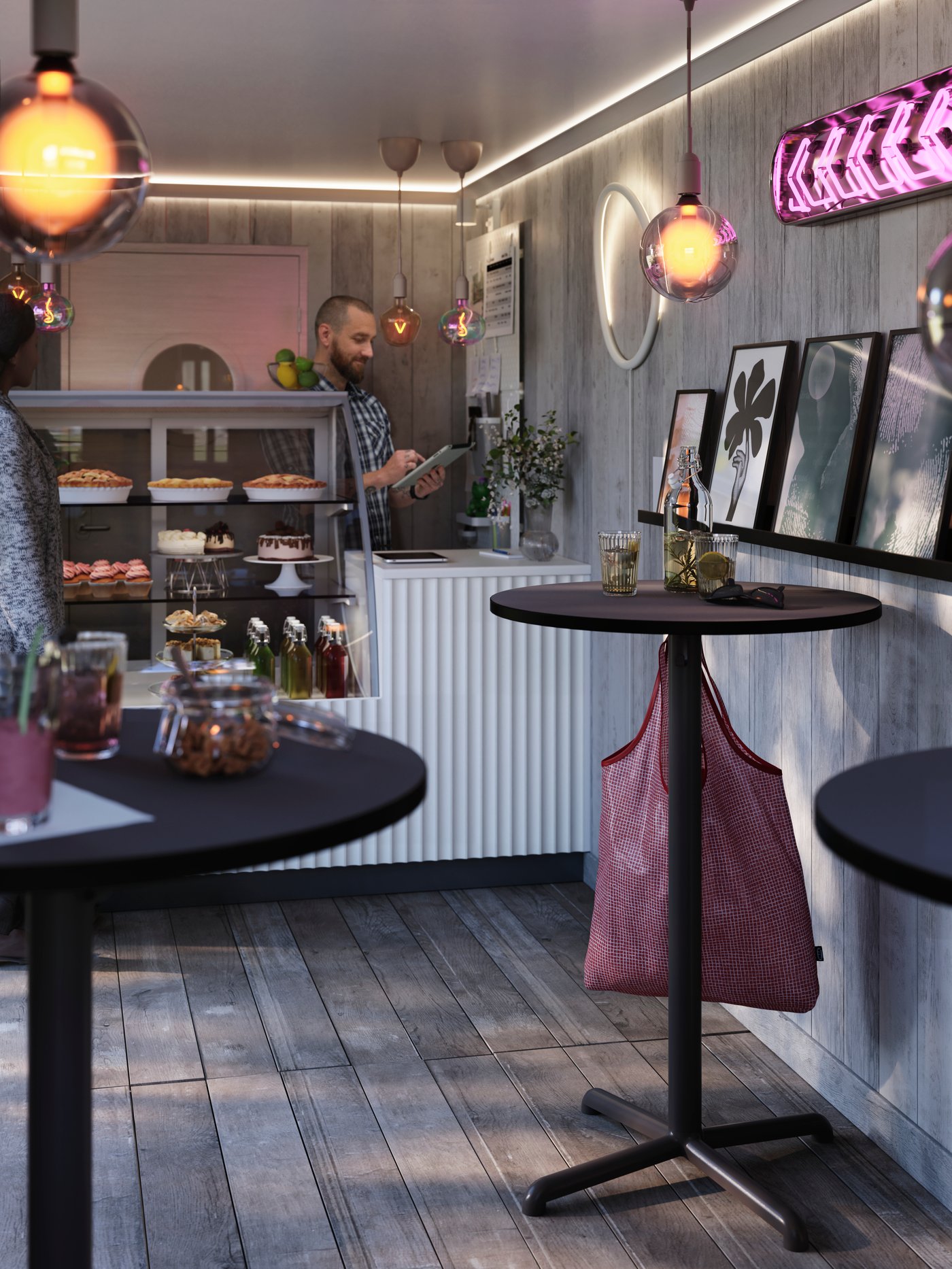 Litet café med runda bord, bakverk i en disk, väggdekor och varma lampor, där en person står bakom disken.