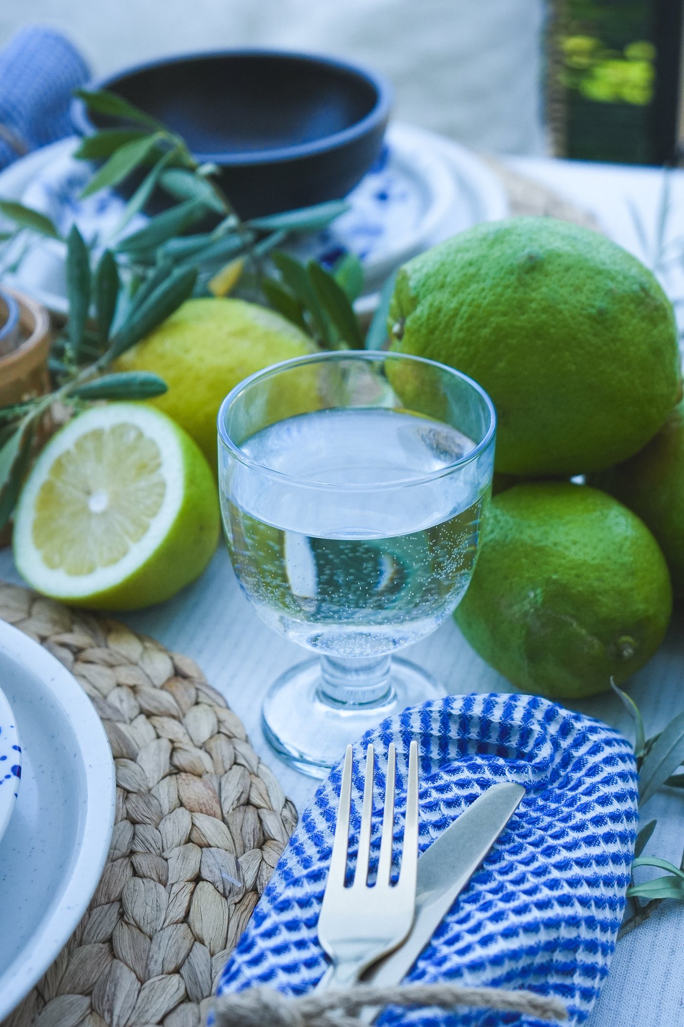Gedeckter Tisch mit einem FRUKTDUVA Glas Wasser, Besteck in blau-weissem Tuch, frischen Limetten und Olivenzweigen als mediterrane Dekoration.