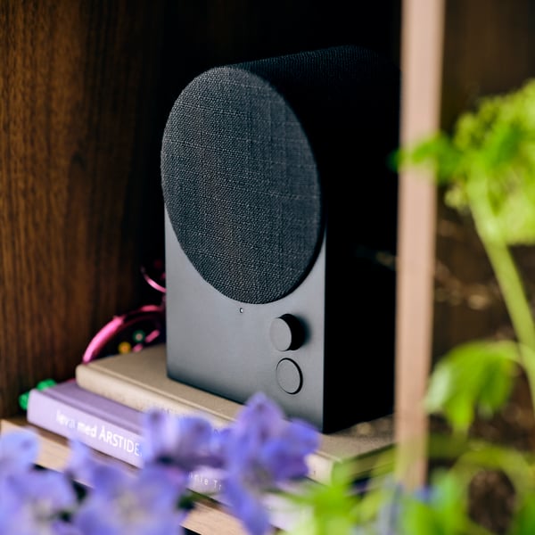 Un mueble de almacenamiento con estantes que sostiene una bocina Bluetooth NATTBAD colocada sobre varios libros y una planta verde.