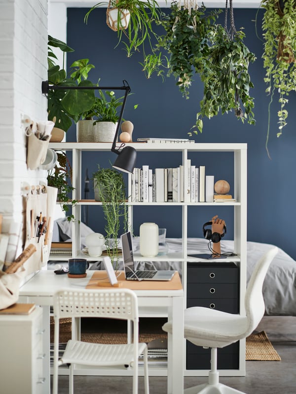 Une table MELLTORP blanche, une chaise blanche, une chaise beige et une lampe murale noire au-dessus, à côté d’un élément de rangement KALLAX blanc.