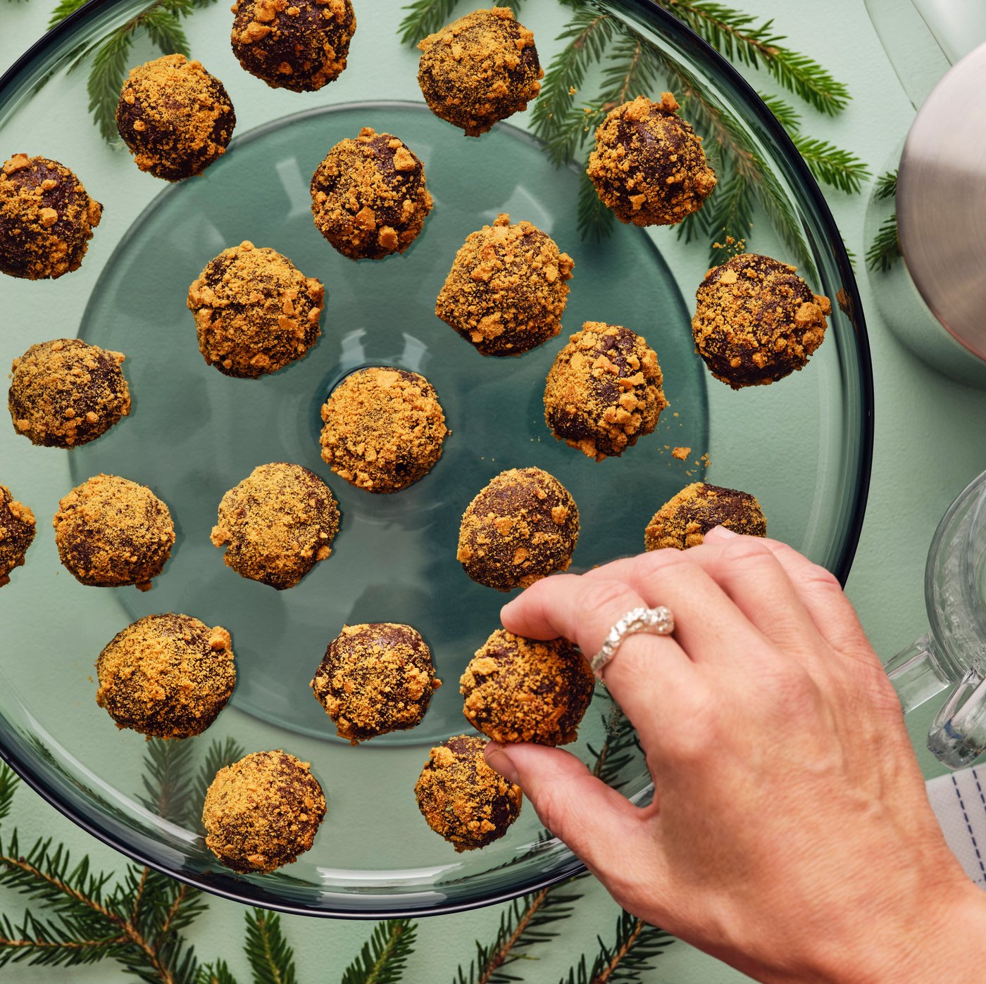 A hand is taking a treat from a glass platter filled with golden brown, crumb-covered truffles, set on a green surface.