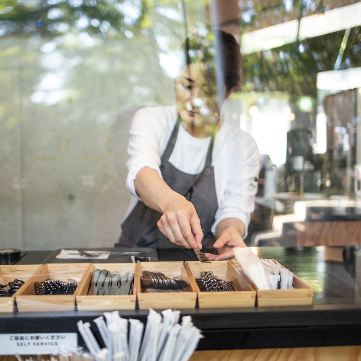 You’re looking through a café window. A woman wearing a white long-sleeve shirt and a grey apron is filling wooden boxes with cutlery.
