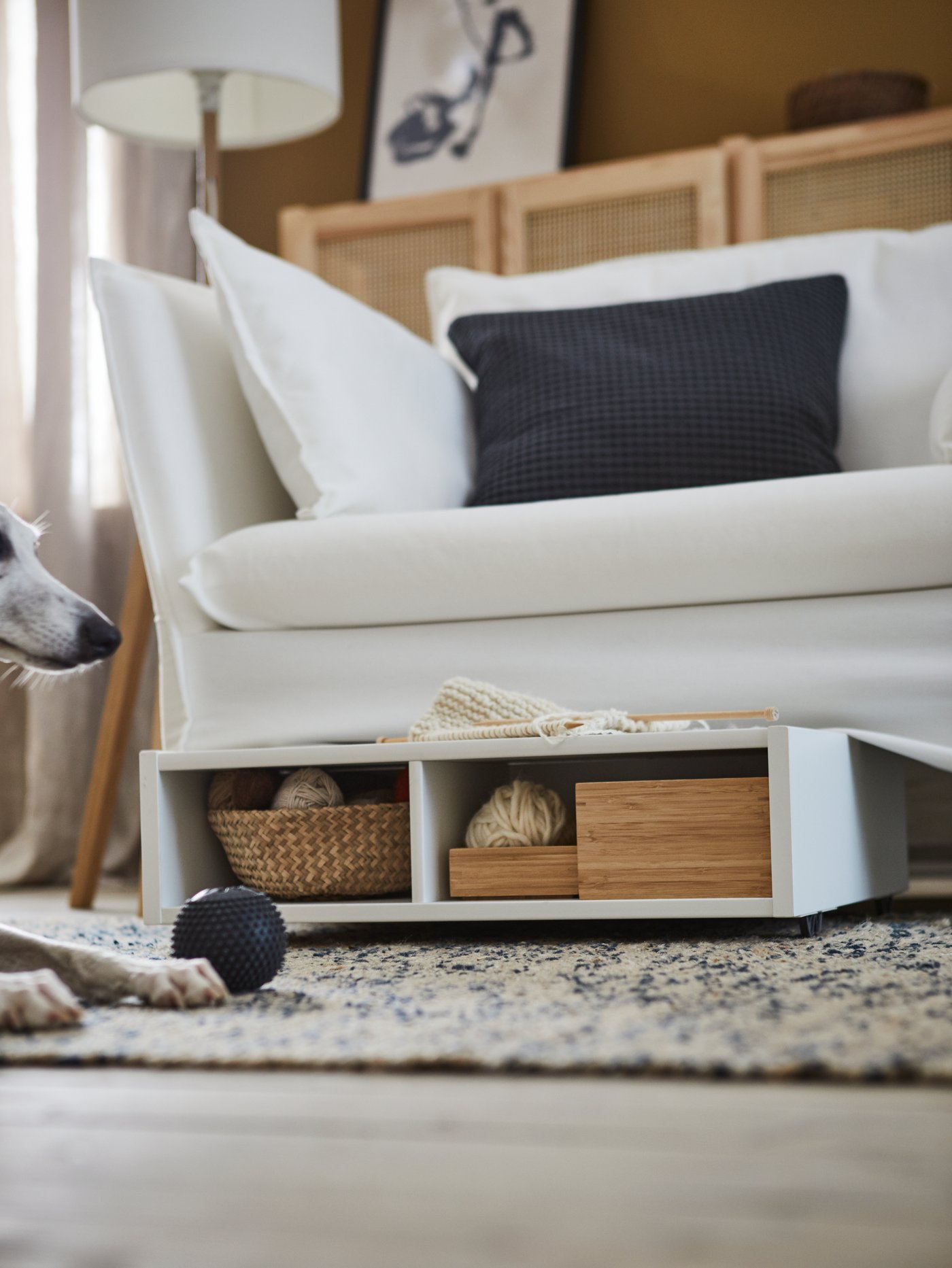 A white underbed storage/bedside table with castors has boxes and a basket inside it under a sofa. A dog with a toy ball is shown on the side.