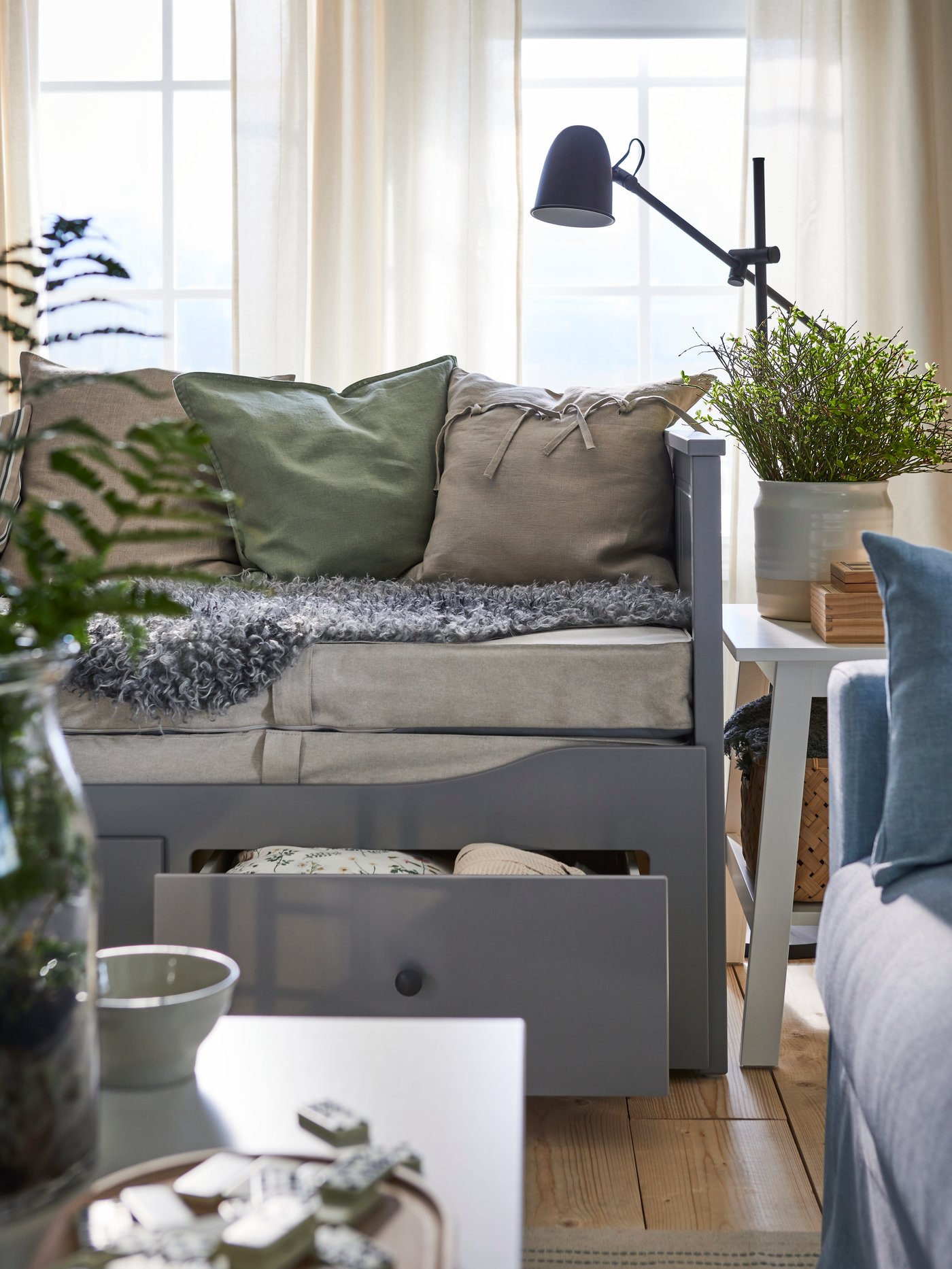 A grey HEMNES day-bed with 3 drawers with one drawer open and plenty of cushions above is in a cottage-style living room.