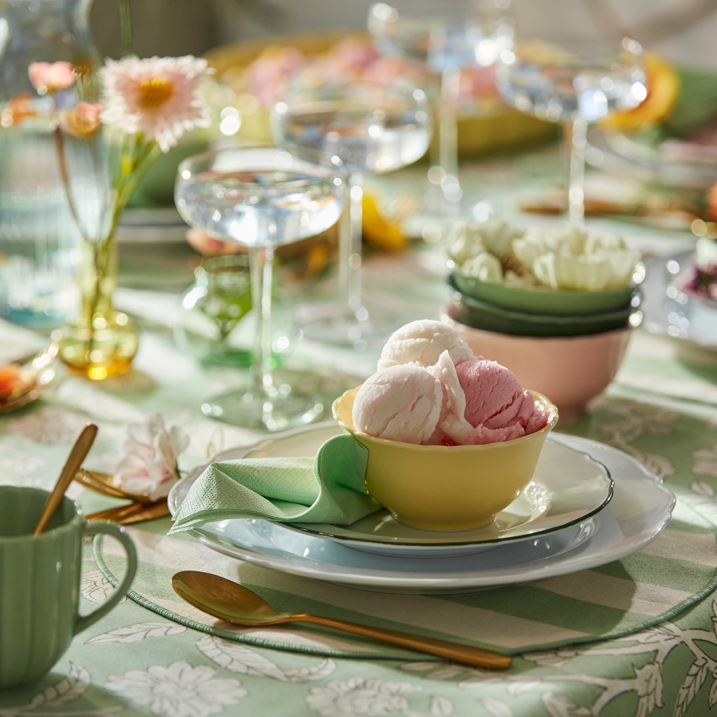 A yellow UPPLAGA bowl holds pink and white ice cream, surrounded by colourful table settings of flowers and glassware.