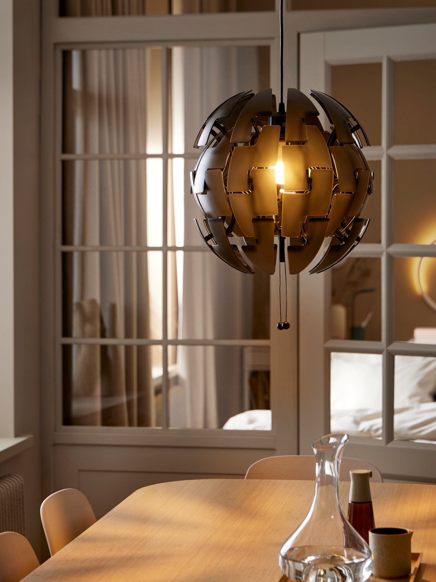 A black IKEA PS 2014 pendant lamp hangs above a VOXLÖV dining table in front of two glass doors. The lamp is lit.