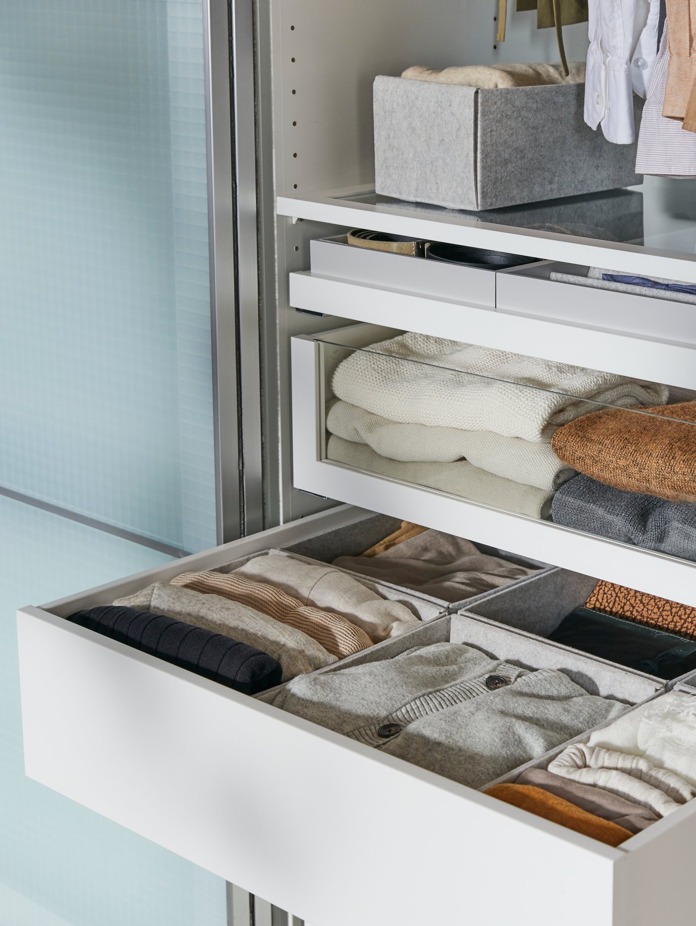 A wardrobe interior has a shelf, a glass-front drawer and an open drawer with sweaters sorted in light grey KOMPLEMENT boxes.