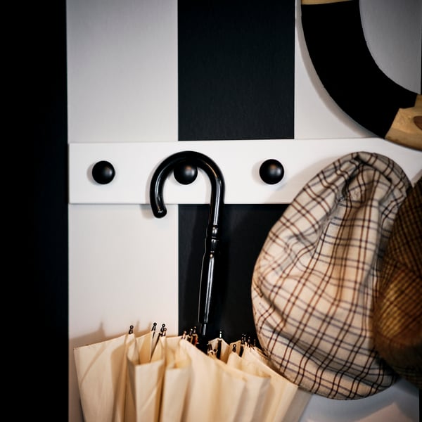Plusieurs articles d'extérieur sont accrochés à des crochets ENERYDA noirs sur une patère LURT blanchie fixée sur un mur à rayures noires et blanches.