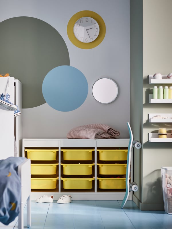A children’s room with a wall clock, a mirror, a skateboard and a white TROFAST storage combination with nine yellow boxes.