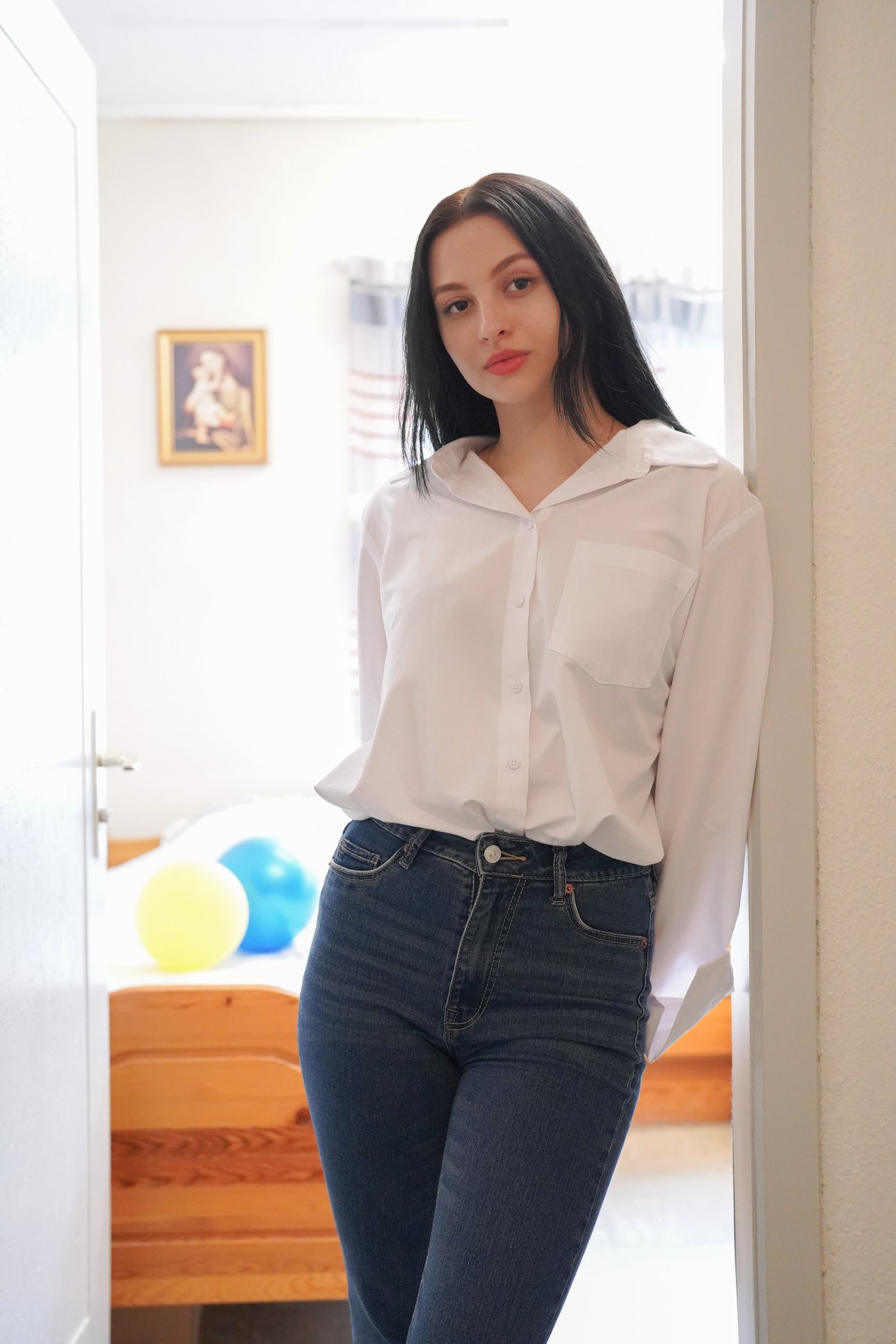 Jeune fille vêtue d’un jeans et d’un haut blanc, adossée à l’encadrement de la porte de son séjour.