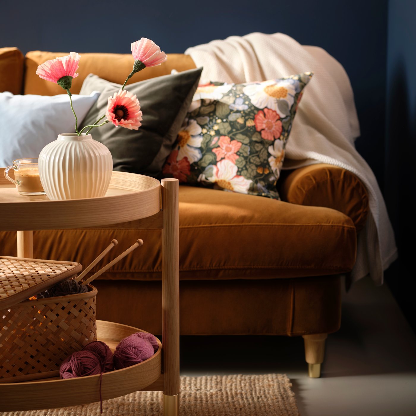 A brown sofa with SANELA and ARENBOK cushion covers and a white HUMLEMOTT throw, by a coffee table with knitting equipment.