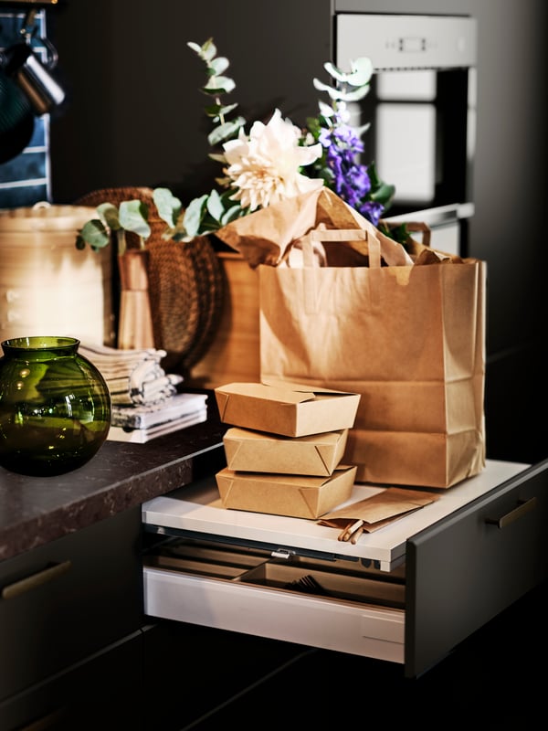 A green KONSTFULL vase on a worktop next to an open UTRUSTA pull-out work surface with a brown paper bag on top.