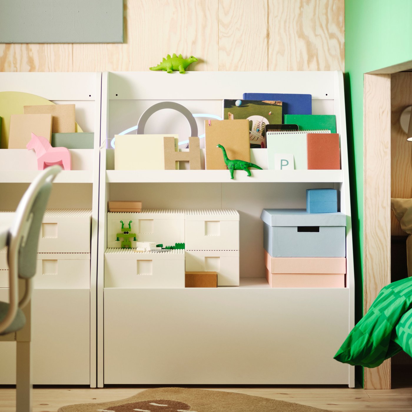 An einer Wand stehen zwei weisse BERGIG-Bücherregale mit Stauraum, in denen verschiedene Kisten, Bücher und Spielsachen untergebracht sind.