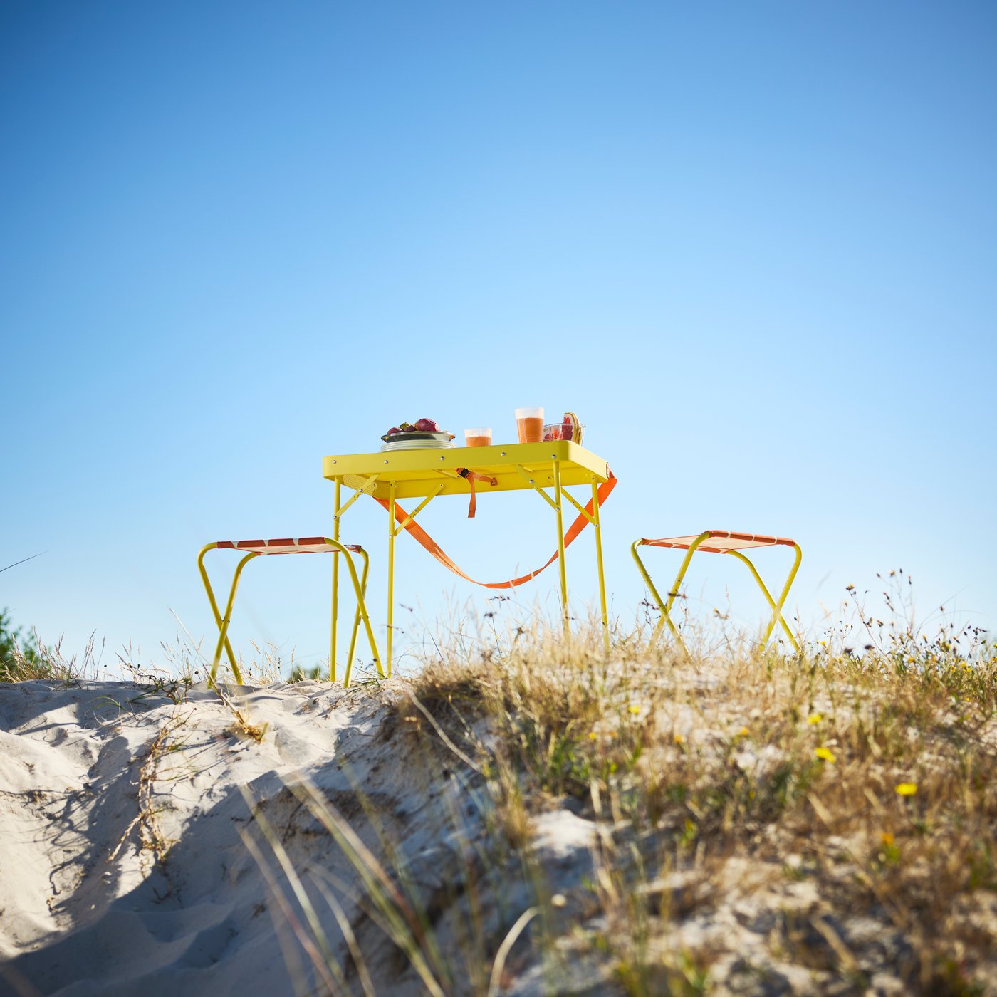 Eine Dünenlandschaft mit einem leuchtend gelben STRANDÖN Klapptisch und zwei dazu passenden Hockern