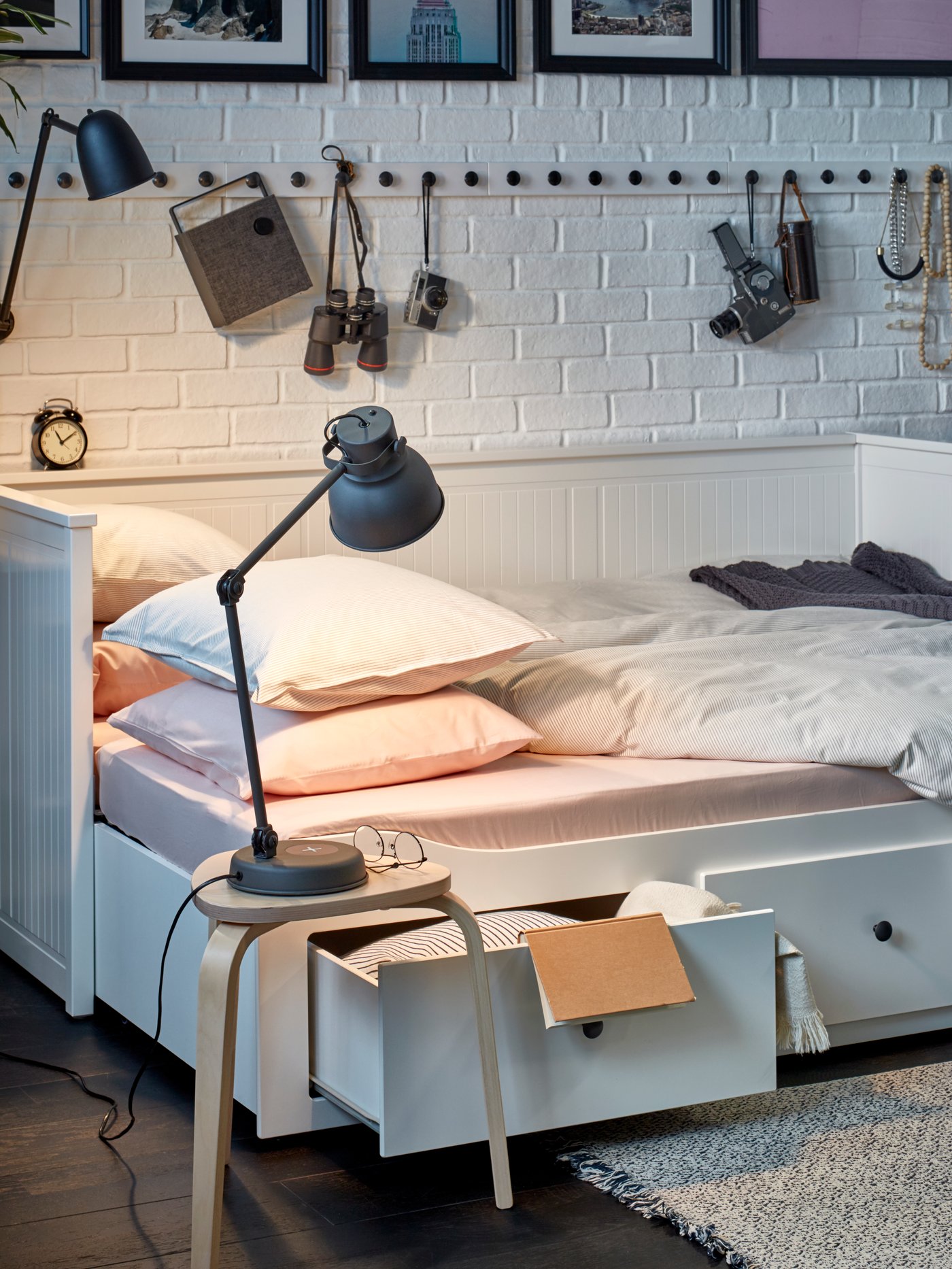 Ein HEMNES Tagesbett in Weiss mit drei Schubladen in seiner Bettfunktion. Neben dem Bett ist ein Hocker mit Leuchte zu sehen.