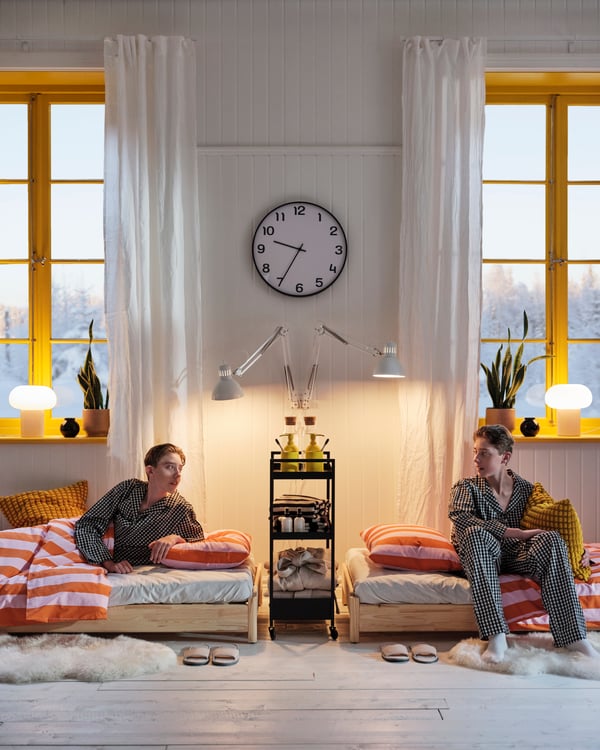 Deux gars sont assis sur un lit dans une pièce avec de grandes fenêtres jaunes, au centre des fenêtres est accrochée une horloge murale PLUTTIS.