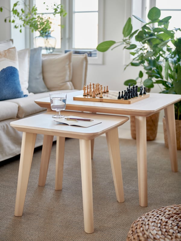 Mysigt vardagsrum med två träbord, ett med ett schackbräde i pågående spel och ett mindre med ett glas vatten och en öppen anteckningsbok. I bakgrunden syns en beige soffa med blå och vita kuddar samt gröna växter vid fönstren.